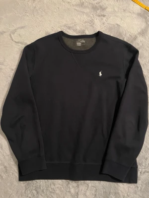 Mörkblå sweatshirt från Polo Ralph Lauren - Klassisk mörkblå sweatshirt från Polo Ralph Lauren med rund halsringning och broderad vit logga på bröstet. Tröjan har ribbade muddar vid ärmslut och nederkant, perfekt för en stilren och avslappnad look.