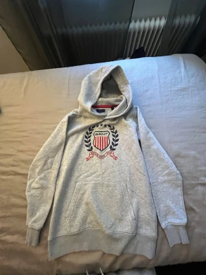 Grå hoodie från Gant  - Snygg grå hoodie från Gant med stort broderat emblem på bröstet i rött, vitt och blått. Hoodien har klassisk känguruficka och ribbade muddar vid ärmar och nederkant. Perfekt för en avslappnad och sportig stil.