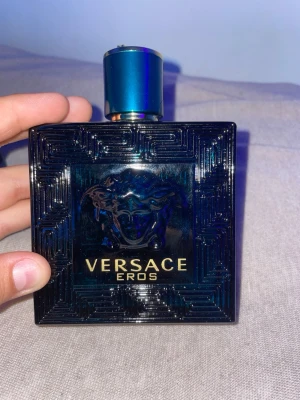 Versace Eros Eau de Toilette 100ml - Versace Eros Eau de Toilette i en lyxig, mörkblå glasflaska med guldtext och Medusa-huvud i relief. Flaskan har en fyrkantig form och ett blått lock. Med tillhörande blå kartong med samma Medusa-detalj och guldtext. Volym: 100 ml.
