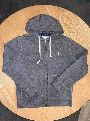 Polo Ralph Lauren Zip Hoodie Mörkgrå – Storlek M - Säljer en Polo Ralph Lauren zip-hoodie i mörkgrå färg.  Klassisk modell med vit Polo-logo på bröstet, huva med vita snören och dragkedja framtill.  Storlek: M Skick: Bra skick. Tvättad och klar.  Unisex: Ja Mått: Kan tas vid förfrågan.