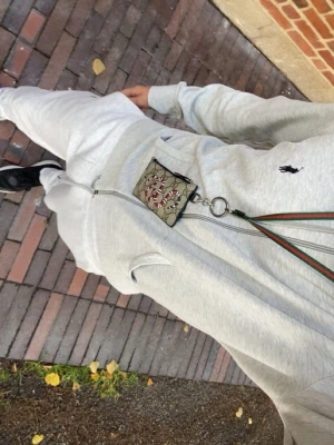 Grå mjukisdress från Polo Ralph Lauren - Säljer en ljusgrå mjukisdress från Polo Ralph Lauren med dragkedja på hoodien och matchande byxor med dragsko i midjan. Setet har den klassiska loggan broderad på både överdel och byxa. Perfekt för chill dagar hemma eller när du vill vara bekväm.