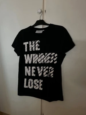 Svart t-shirt med vit text  - Cool svart t-shirt med stor vit text på framsidan där det står 'THE WINNER NEVER LOSE'. Klassisk rund hals och korta ärmar. Perfekt för dig som gillar statement-plagg och enkel stil. Denimday