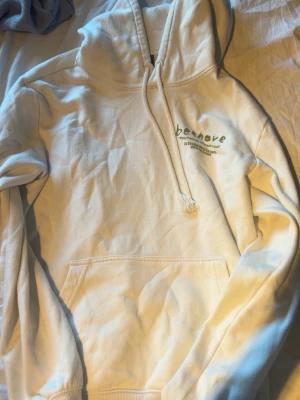 Vit hoodie med grön text och tryck från HM - Vit hoodie med huva och dragsnören. På bröstet står det 'beehave' i grön text och på ryggen finns ett stort grönt tryck med blommor och bin samt texten 'Tiny heroes, colossal impact'. Stor magficka framtill och ribbade muddar. Passar perfekt nu till höst🍂