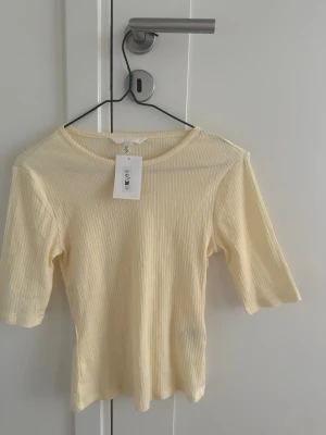 Ljusgul ribbad topp från H&M - Säljer en ljusgul ribbad topp från H&M med rund halsringning och halvlånga ärmar. Toppen är i ett mjukt, stretchigt material och har en smickrande, figurnära passform. Perfekt att styla med jeans eller kjol för en fräsch look.