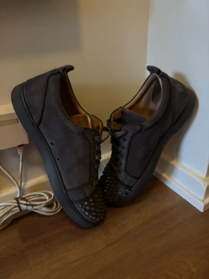 Svarta sneakers med nitar. Christian Louboutin  - Säljer ett par svarta sneakers i mocka med coola nitar på tån och klassisk röd sula. Skorna har lågt skaft och snörning framtill. Insidan är fodrad i beige läder för extra komfort. Perfekta för dig som vill sticka ut. Dom har lite skrapor på några av nitarna men det är inget som märks jätte tydligt när man går med dom.