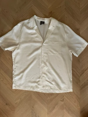 Vit kortärmad skjorta med öppen krage - Stilren vit skjorta med korta ärmar och öppen krage. Skjortan har en bröstficka och knäppning framtill. Perfekt till sommaren och tillverkad i ett mjukt, lätt material som ger en avslappnad look.