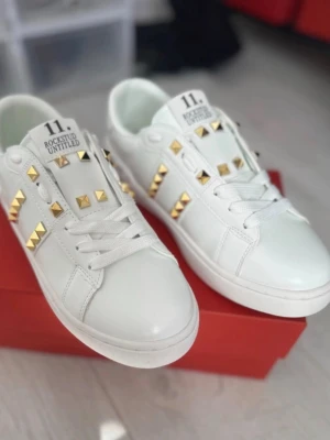 Vita sneakers med guldfärgade nitar - Snygga vita sneakers med guldfärgade nitar i rutmönster. Skorna har klassisk låg profil, vita snören och detaljer i vitt läder. Märket 'Rockstud Untitled' syns på plösen. Perfekta för dig som vill sticka ut med coola detaljer.