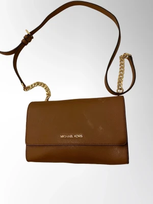 Michael Kors Brun axelremsväska  - Snygg brun handväska från Michael Kors med guldfärgad kedjedetalj på axelremmen. Väskan har en stilren, rektangulär form och är tillverkad i läder. Perfekt för dig som vill ha en trendig och klassisk accessoar.