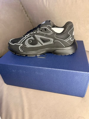 Svarta dior B30 sneakers - Snygga svarta sneakers med vita kontrastdetaljer och grov sula. Ovandelen är i mesh och syntetmaterial med coola mönster och reflekterande linjer. Skon har en sportig look och passar perfekt till streetwear-stilen.