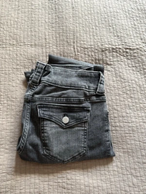 Grå jeans med fickdetaljer - Säljer ett par grå jeans med snygga fickdetaljer bak och dubbla knappar fram. Jeansen har klassisk femficksmodell och bootcut. Materialet är stretchigt denim som sitter skönt och följer kroppen. Storlek 170 i barn men passar alla som är upp till 156 cm långa då jag har sytt upp dom så de passar mig som är 156cm.