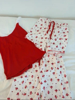 Röd och vit blommig pyjamas-set - Sött pyjamas-set med ett rött linne med vita spetsdetaljer och matchande vita byxor och skjorta med röda blommor. Linnet har tunna axelband och ribbad struktur, medan byxorna och skjortan har ett romantiskt blommönster och röda detaljer. Perfekt för mysiga kvällar.