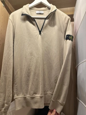 Beige halfzip från Stone Island - Snygg beige halfzip-tröja från Stone Island med svart logopatch på ärmen. Tröjan har hög krage med dragkedja och långa ärmar. Perfekt för en avslappnad och trendig stil.
