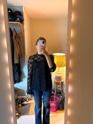 Svart spetsblus med hög krage - Säljer en svart blus i genombruten spets med hög krage och långa ärmar. Blusen har ett elegant mönster och är lätt transparent, perfekt för dig som gillar en trendig och stilren look. Passar till både jeans och kjol. 