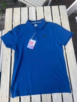 Ny Helly Hansen Pikétröja | Oanvänd med lappar kvar - Säljer en helt ny och oanvänd blå pikétröja från Helly Hansen med originallapparna kvar. Perfekt passform och hög kvalitet. Klassisk modell som passar både till vardags och aktiva dagar. 