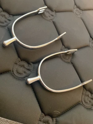 Silvriga sporrar i metall - Ett par klassiska silvriga sporrar i metall, perfekta för ridning. De har en enkel och stilren design med rundad form och öppna ändar för remmar. Passar till de flesta ridstövlar och ger bra kontroll vid ridning.