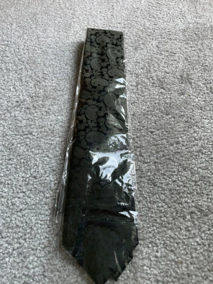 Svart slips med paisleymönster - Snygg svart slips med diskret paisleymönster som ger en elegant touch till din outfit. Slipsen är tillverkad i ett glansigt tyg och levereras i plastförpackning. Perfekt för dig som vill sticka ut lite extra med detaljer.