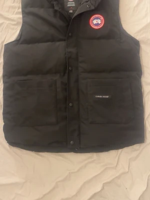 Svart dunväst från Canada Goose - Snygg svart dunväst från Canada Goose med klassisk logga på bröstet och två stora fickor framtill. Västen har tryckknappar och dragkedja samt diskret Canada Goose-märke på ena fickan. Perfekt för lager-på-lager och håller dig varm.