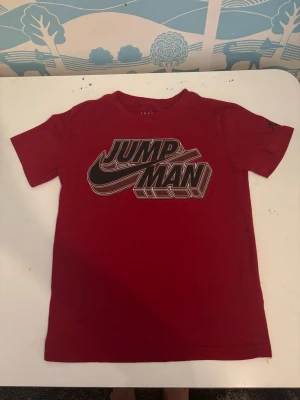 Röd Jordan Jumpman t-shirt - Snygg röd t-shirt från Jordan med stort svart och vitt Jumpman-tryck på bröstet. Tillverkad i en mjuk bomulls- och polyestermix, perfekt för en sportig och avslappnad stil. Klassisk passform med rund hals och korta ärmar.