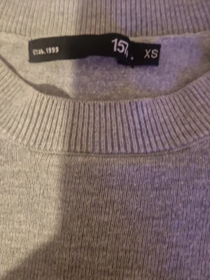 Ljusgrå tröja från 157, XS - En stilren ljusgrå tröja från 157 i storlek XS. Tröjan har rund halsringning, långa ärmar och ribbade muddar vid ärmslut och nederkant. Perfekt för en enkel och clean look.