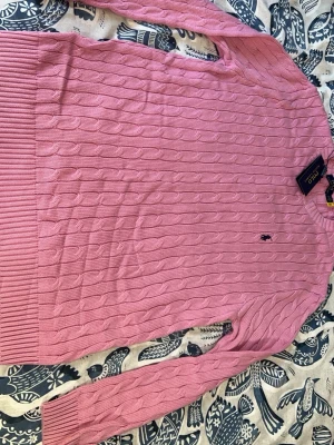 Rosa kabelstickad tröja från Polo Ralph Lauren - Kabelstickad rosa tröja från Polo Ralph Lauren med klassisk rund halsringning och svart broderad logga på bröstet. Tröjan har långa ärmar och breda ribbade muddar vid ärmslut och nederkant. Perfekt för en stilren och avslappnad look.