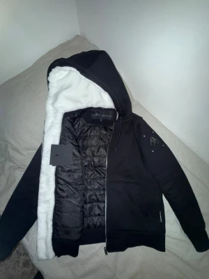 Svart teddyjacka med huva från Moose Knuckles - Svart teddyjacka från Moose Knuckles med vit fluffig teddyfoder i huvan och quiltad insida. Jackan har dragkedja, snörning i huvan och en logga på ärmen. Perfekt för kyliga dagar och riktigt snygg streetstil.