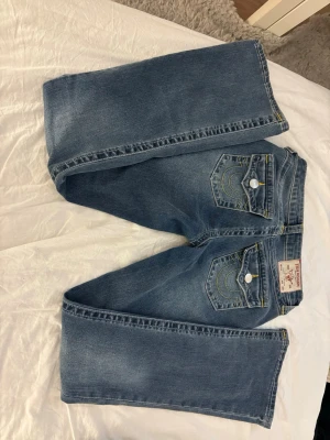 True religon jeans - Säljer ett par klassiska blå bootcut jeans  med kontrastsömmar och snygga detaljer på bakfickorna. Jeansen har fem fickor, vita knappar och etikett bak. Perfekta för en avslappnad och trendig look.
