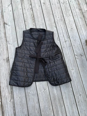 Svart quiltad dunväst med knytband - Stilren svart dunväst med quiltad design och två breda knytband framtill. Västen är ärmlös, har rund halsringning och lätt glansigt material. Perfekt för lager på lager och ger en modern touch till din outfit.