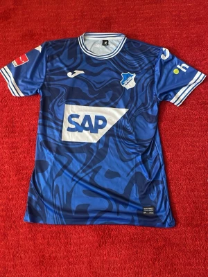 Blå Hoffenheim matchtröja Joma - Säljer en blå matchtröja från Hoffenheim med mönstrad design och vita detaljer. Tröjan har klubbmärke på bröstet, SAP-logga framtill och tryck på ärmarna. Tillverkad i lätt och ventilerande material, perfekt för fotboll eller träning.