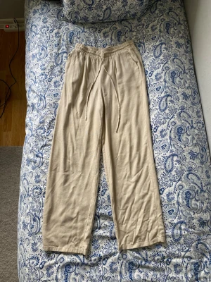 Beiga vida linnebyxor  - Säljer ett par vida beiga linnebyxor med avslappnad passform. Byxorna har resår i midjan och perfekt mått på snörena. Perfekta för en chill och stilren look under varma dagar. Använt ett fåtal gånger.