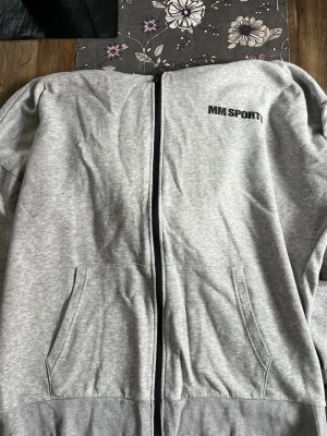 Grå zip hoodie från MM Sports - Säljer en grå hoodie med dragkedja från MM Sports. Tröjan har fickor framtill och ribbade muddar vid ärmslut och nederkant. Perfekt för chill dagar eller gymmet. Materialet är mjukt och bekvämt, troligen bomullsblandning. Diskret logga på bröstet.