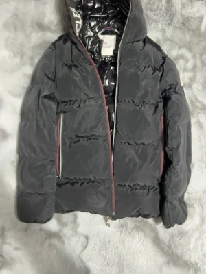 Svart dunjacka från Moncler - Snygg svart dunjacka från Moncler med huva och glansigt innerfoder. Jackan har dragkedja med röda detaljer, två fickor med dragkedja och stora Moncler-loggor på insidan. Perfekt för kalla vinterdagar och riktigt trendig look.