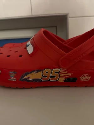 Röda Crocs Cars Lightning McQueen - Säljer ett par röda Crocs med Lightning McQueen-motiv från Disney Cars. Skorna har perforerad ovandel, justerbar hälrem och är tillverkade i lätt och bekvämt gummimaterial. Dekorativa detaljer som ögon och nummer 95 på sidorna. 
