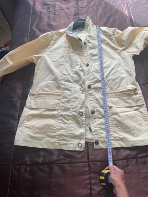 Vintage Emm ljusgul jacka, storlek 46 - Snygg ljusgul vindjacka från Emm med dragkedja och tryckknappar framtill. Jackan har två stora fickor med lock och en hög krage. Tillverkad i ett lätt och vindtätt material, jag små axelvaddar 