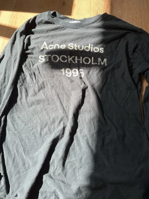 Svart långärmad t-shirt Acne Studios - Säljer en svart långärmad t-shirt från Acne Studios med tryck 'Acne Studios STOCKHOLM 1996' på bröstet. Tillverkad i mjuk bomull med en klassisk passform och rund hals. Perfekt för dig som gillar stilrena och tidlösa plagg. Stor i storlek 