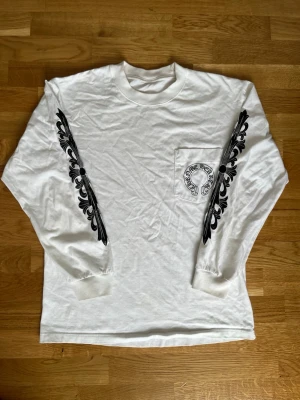 Vit långärmad topp Chrome Hearts - Vit långärmad topp från Chrome Hearts med svarta tryck längs ärmarna och stor logga på ryggen. Bröstficka med tryck och rund halsringning. Perfekt för dig som gillar streetwear och unika detaljer.