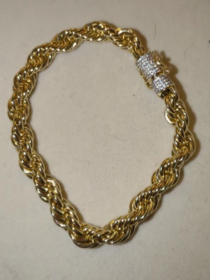 Guld cordell med diamantlås  - Snyggt armband i guldfärgad metall med flätad design. Spännet är dekorerat med små gnistrande stenar som ger extra bling. Perfekt accessoar för dig som gillar att sticka ut med detaljer.