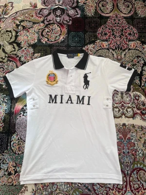 Vit piké med svart krage från Polo Ralph Lauren - Vit pikétröja från Polo Ralph Lauren med svart krage och detaljer. Stor broderad logga på bröstet, texten MIAMI och ett snyggt klubbmärke. Klassisk passform och korta ärmar, perfekt till en avslappnad stil.
