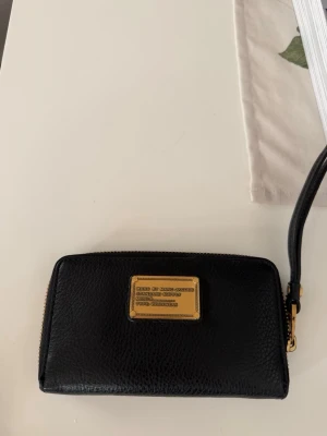 Svart plånbok Marc by Marc Jacobs - Snygg svart plånbok i äkta läder från Marc by Marc Jacobs. Plånboken har guldfärgade detaljer, dragkedja runtom och flera fack för kort och mynt. Klassisk design med logoplatta framtill och handledsrem för enkel användning.