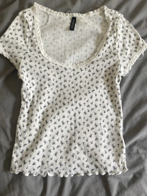 Vit blommig ribbad topp - Söt vit ribbad topp med små blommor i svart. Toppen har korta ärmar, rundad urringning och vågig kant nertill och vid ärmslut. Perfekt för dig som gillar en feminin och avslappnad stil.