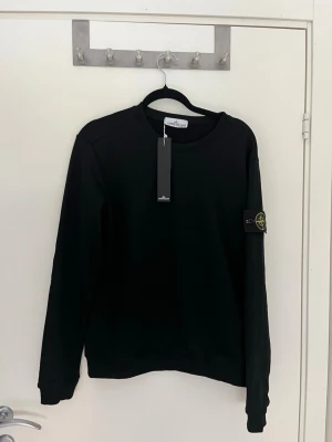 Svart sweatshirt från Stone Island - Svart sweatshirt från Stone Island med klassisk rund halsringning och ikonisk logotyp-patch på vänster ärm. Tröjan har ribbade muddar vid ärmslut och nederkant. Perfekt för en clean och stilren look. Den är M men passar mer som S