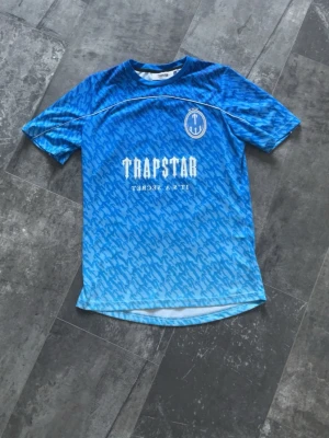 Trapstar T-shirt  - Säljer en helt ny Trapstar tröja med vit logga och text på bröstet samt stort nummer 22 på ryggen. Tröjan har ett mönster i olika blå nyanser, rund halsringning och korta ärmar. Perfekt för träning eller streetwear.