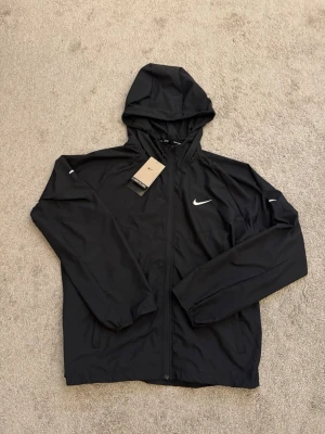 Svart Nike Miller vindjacka  - Snygg svart löparjacka från Nike med huva och dragkedja framtill. Jackan har justerbar dragsko i huvan, elastiska muddar och en vit Nike-logga på bröstet. Tillverkad i lätt och funktionellt material, perfekt för löpning och träning utomhus.