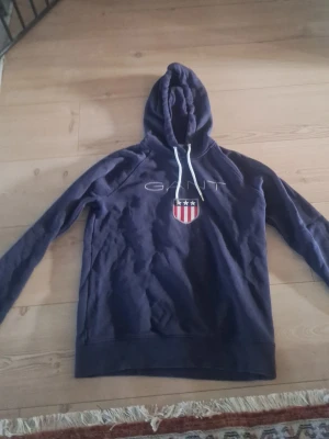Mörkblå hoodie från Gant - Snygg mörkblå hoodie från Gant med vit dragsko i huvan och ett broderat Gant-logotyp samt ett sköldmärke med stjärnor och ränder på bröstet. Klassisk passform och mjukt material, perfekt för en avslappnad stil.