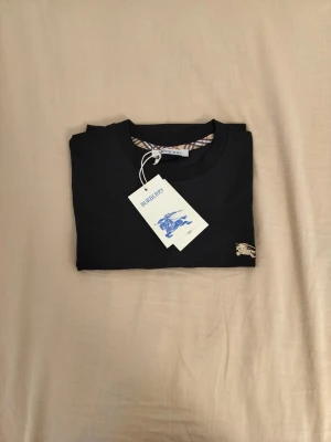 Svart Burberry t-shirt i bomull - Snygg svart t-shirt från Burberry med klassisk rund hals och diskret Burberry-logga i guld på bröstet. Tillverkad i mjuk bomull och har korta ärmar. Perfekt basplagg med exklusiv känsla och stilren design.