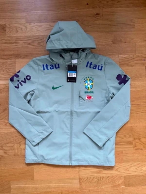 Ljusgrön Brasilien vindjacka Nike - Snygg ljusgrön vindjacka från Nike med Brasilien-logga och sponsormärken som Itau och Vivo. Jackan har huva, hel dragkedja och meshfoder på insidan. Perfekt för dig som gillar sportig stil och vill sticka ut med landslagskänsla.