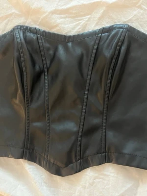 Svart korsett i skinnimitation - Stilren svart korsett i skinnimitation med markerade sömmar framtill. Axelbandslös modell som ger en edgy och trendig look. Perfekt att styla med höga byxor eller kjol för en cool outfit.