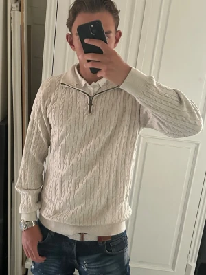 Kabelstickad tröja halvzip  - Snygg beige kabelstickad tröja med halvzip och hög krage. Tröjan har ribbade muddar vid ärmslut och nederkant samt ett klassiskt kabelmönster. Perfekt för lager på lager och passar till jeans eller chinos.