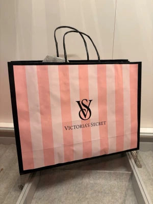 Shoppingväska Victoria's Secret - Stor shoppingväska från Victoria's Secret i klassiskt rosa och ljusrosa randigt mönster med svarta handtag och svart ram. Tillverkad i papper och har loggan tryckt på framsidan. Perfekt för shoppingrundan eller som presentpåse.