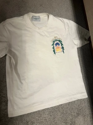 Vit t-shirt  Casablanca - Vit t-shirt från Casablanca med tryck fram och bak. På bröstet finns ett färgglatt motiv med texten 'Avenida Casablanca' och på ryggen ett större tryck av en byggnad inramad av gröna blad. T-shirten har rund hals och korta ärmar, tillverkad i mjuk bomull.