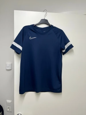 Mörkblå Nike Dri-FIT sporttröja - Snygg mörkblå sporttröja från Nike med vita detaljer på ärmarna och klassisk Nike-logga på bröstet. Tillverkad i Dri-FIT-material som andas och håller dig torr under träningen. Perfekt för fotboll, gym eller löpning.